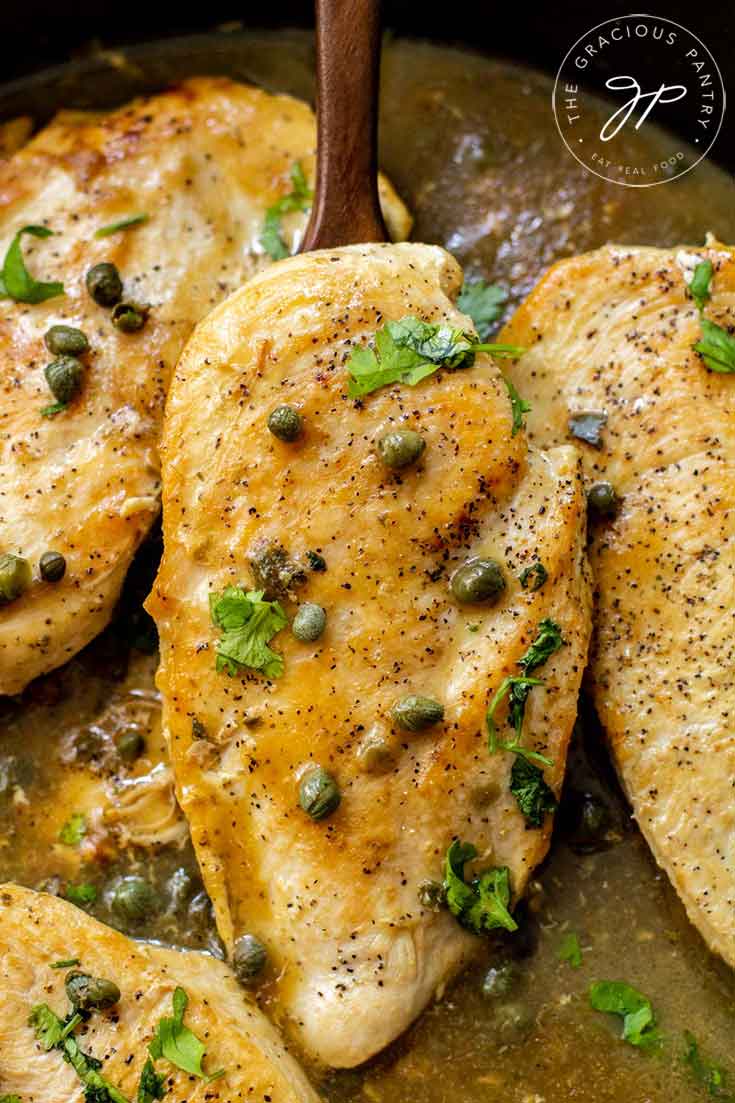 And up close view of this lemon caper chicken still sitting in the skillet with a serving spoon lifting up one of the chicken breasts. It has capers sprinkled over it as well as fresh parsley.