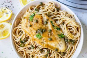 A single chicken breasts shown served over some whole grain pasta. Cut lemons sit to the side of the white bowl.