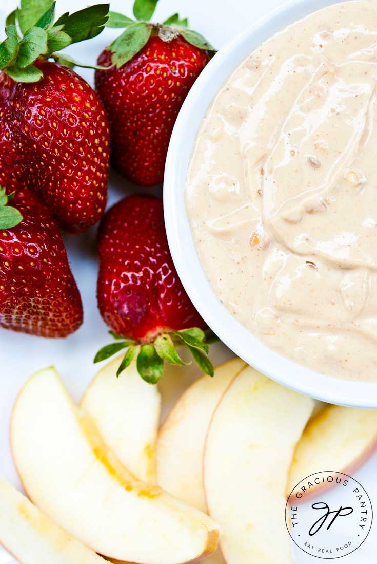 A close up shot of this Clean Eating Peanut Butter Fruit Dip in a white bowl. Only half the bowl is shown in the upper right hand corner. The bowl is surrounded by strawberries and apple slices.