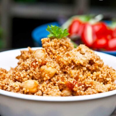 A bowl sits on a wooden table filled with this Indian CousCous Recipe. Fresh tomatoes sit in the background.