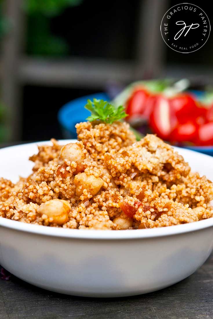 A bowl sits on a wooden table filled with this Indian CousCous Recipe. Fresh tomatoes sit in the background.