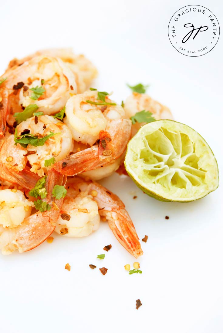 Clean Eating Garlic Lime Shrimp on a white background with a few red pepper flakes sprinkled over them. A half lime that has been squeezed sits next to the shrimp.