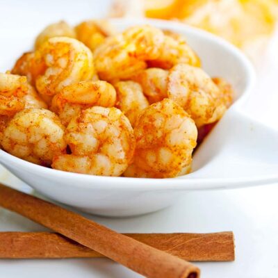 A bowl of bright, orange shrimp sits next to a peeled orange and some cinnamon sticks in this Spicy Orange Shrimp Recipe