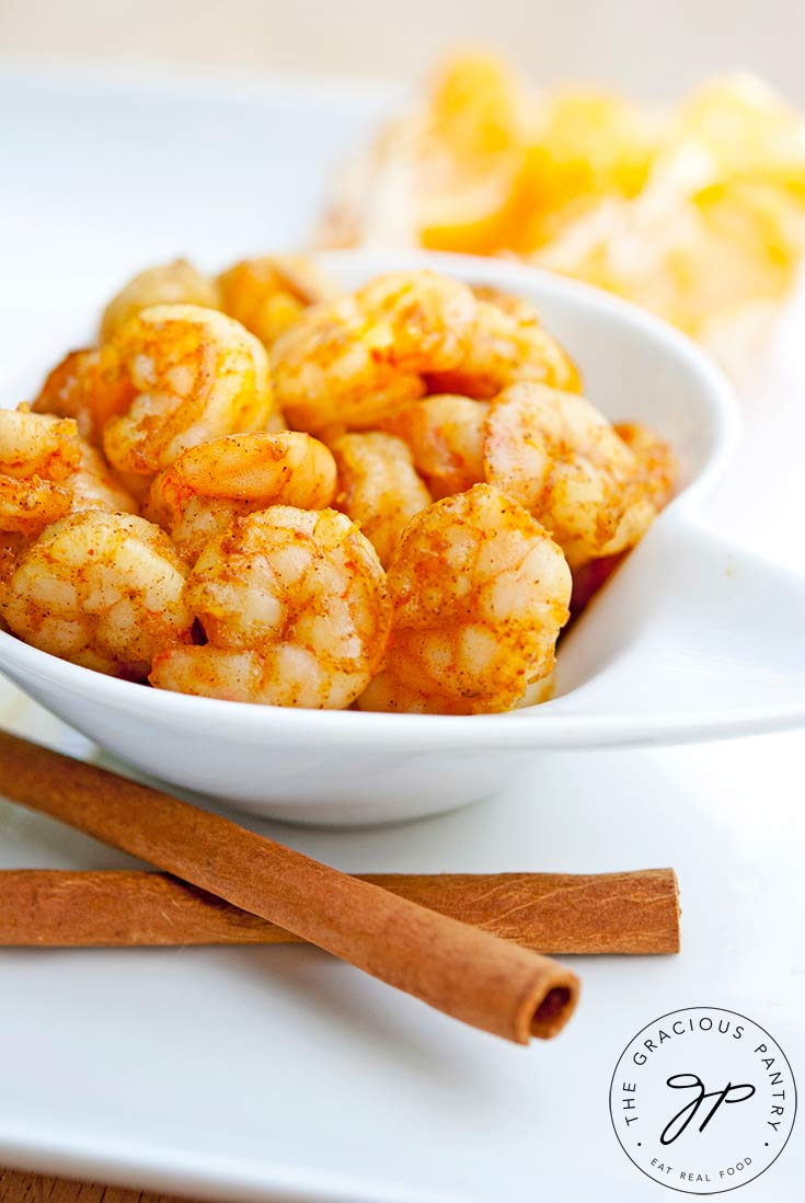 A bowl of bright, orange shrimp sits next to a peeled orange and some cinnamon sticks in this Spicy Orange Shrimp Recipe