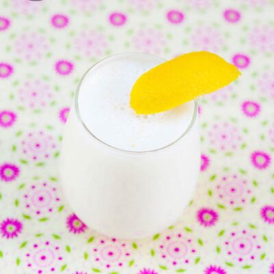 A single glass of this Lemon Smoothie Recipe sits on a flowery tablecloth and has a lemon wedged garnishing the edge of the glass.