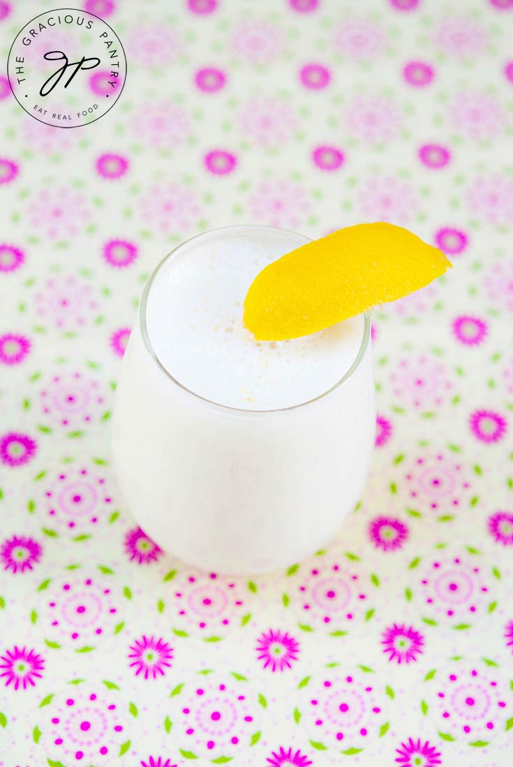 A single glass of this Lemon Smoothie Recipe sits on a flowery tablecloth and has a lemon wedged garnishing the edge of the glass.