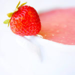 A white bowl sits filled with this delicious, clean eating strawberry soup. A single fresh strawberry sits perched on the handle of the bowl against a completely white background.