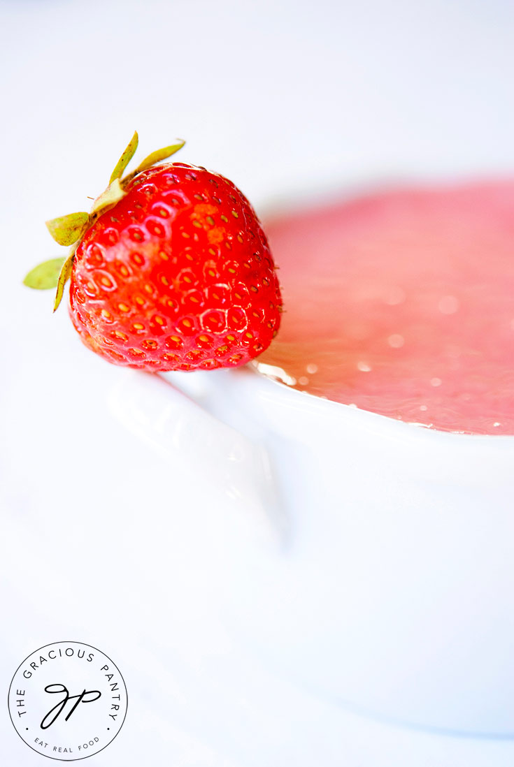 A white bowl sits filled with this delicious strawberry soup. A single fresh strawberry sits perched on the handle of the bowl against a completely white background.