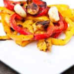 A white plate holds some red and yellow bell peppers with garlic cloves sitting on top from this Clean Eating Barbecued Bell Pepper Recipe.