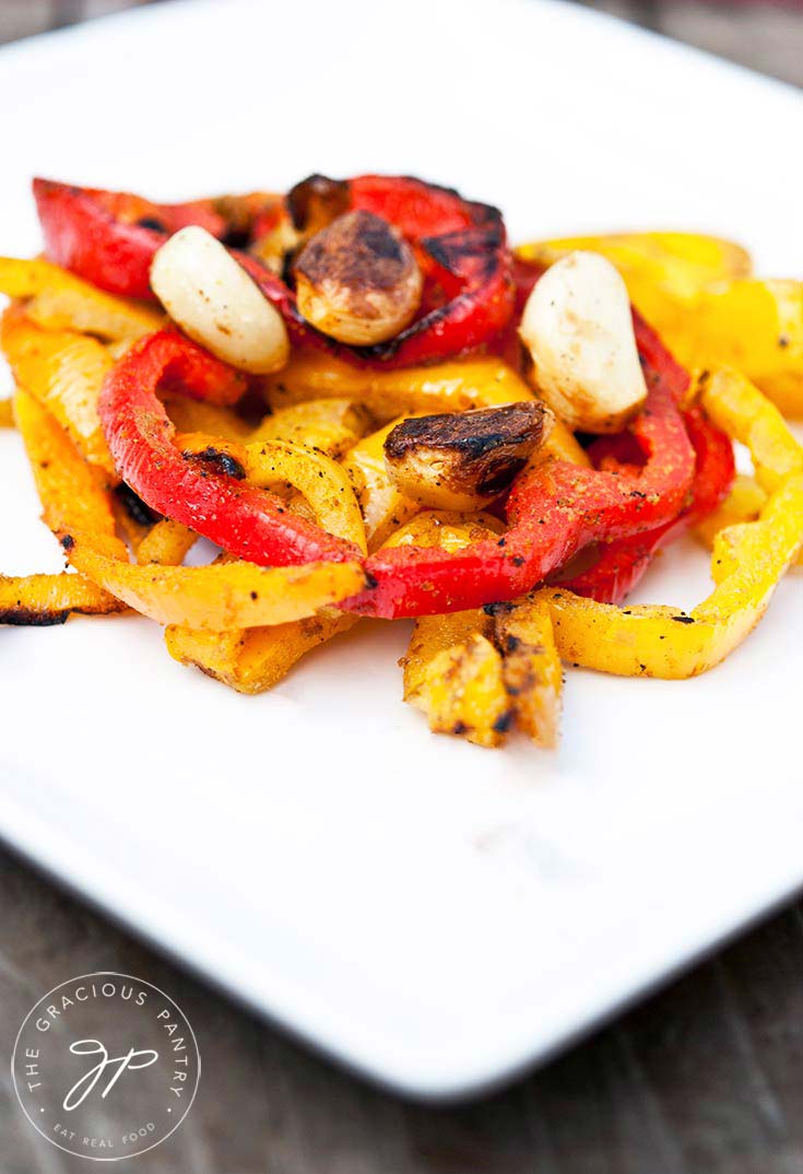 A white plate holds some red and yellow bell peppers with garlic cloves sitting on top from this Clean Eating Barbecued Bell Pepper Recipe.