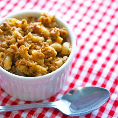 A white bowl sits on a red and white checkered table cloth, filled with this Chili Mac Recipe. A spoon sits beside the bowl.