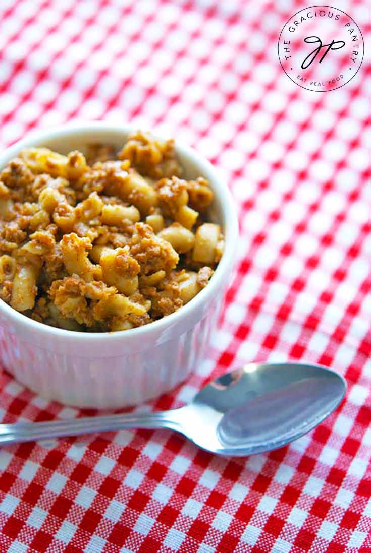 A white bowl sits on a red and white checkered table cloth, filled with this Chili Mac Recipe. A spoon sits beside the bowl.