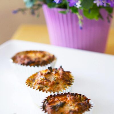 Three of these Spinach And Feta Breakfast Muffins are lined up on a white platter with a beautiful, purple, potted plant sitting next to it.