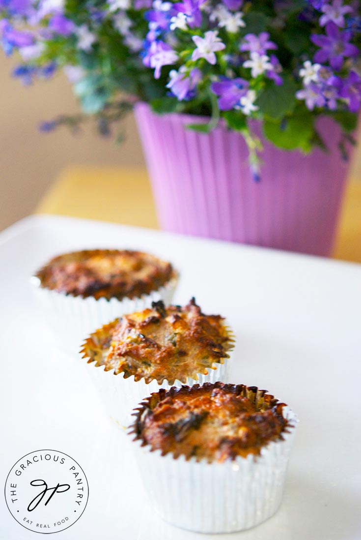 Three of these Spinach And Feta Breakfast Muffins are lined up on a white platter with a beautiful, purple, potted plant sitting next to it. 