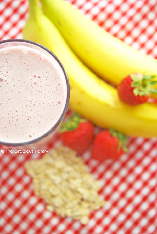 Clean Eating Strawberry Oatmeal Smoothie Recipe shown in a glass on a red and white checkered table cloth. It has bananas, strawberries and oats sitting on the table around the glass.