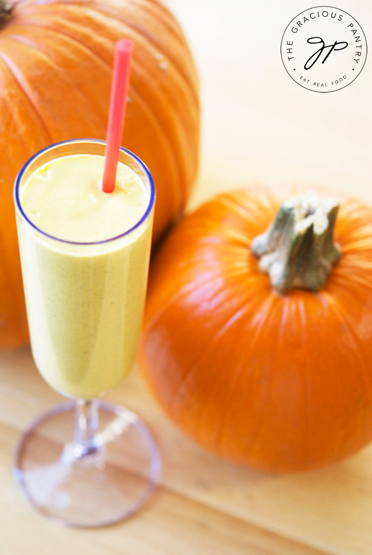 A single champagne glass filled with this Clean Eating Pumpkin Pie Oatmeal Smoothie sits between two pumpkins.