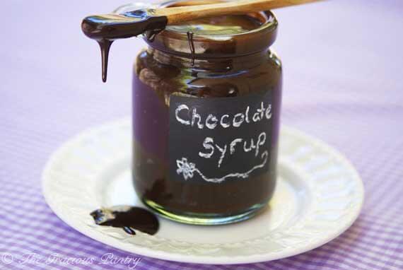 Clean Eating Chocolate Syrup displayed in a clear jar with a small wooden spoon resting on top of the open jar. The spoon has chocolate sauce dripping off of it onto a white plate beneath the jar.