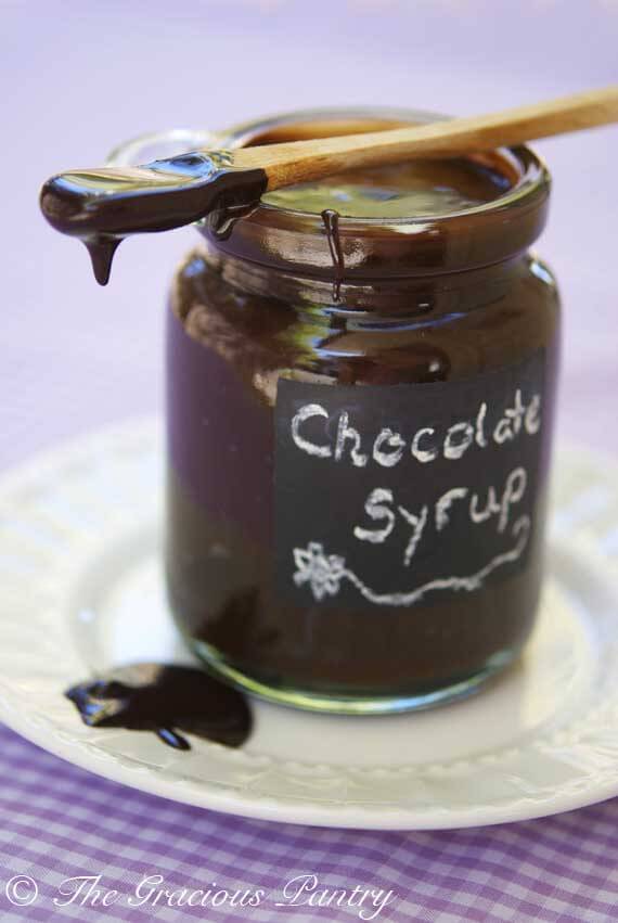 A clear jar with a chalkboard label sits on a plate without a lid. It has a small wooden spoon with the tip covered in chocolate, laying across the top of the jar filled with this Clean Eating Chocolate Syrup