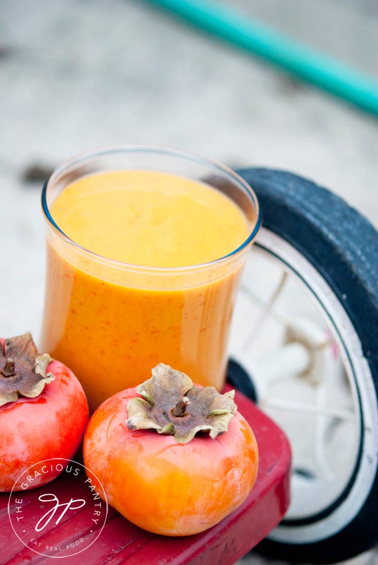 A glass of Clean Eating Kid's Persimmon Smoothie sits on the back step of a red tricycle with two persimmons sitting in front of the glass.