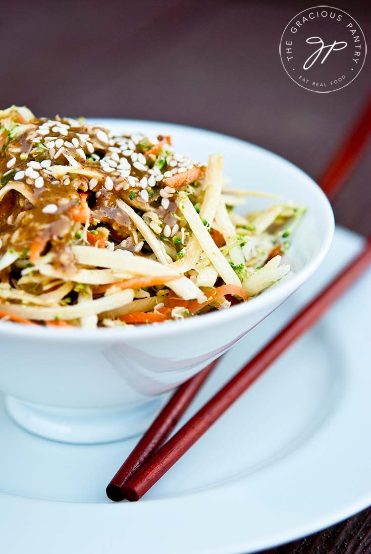 A white bowl filled with Clean Eating Raw Asian Broccoli Salad and topped with brown soy dressing and sesame seeds sits on a brown background with chopsticks resting next to the bowl.