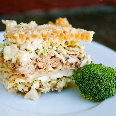 A slice of they Clean Eating White Lasagna Recipe sits on a white plate with a single broccoli floret beside it for garnish.