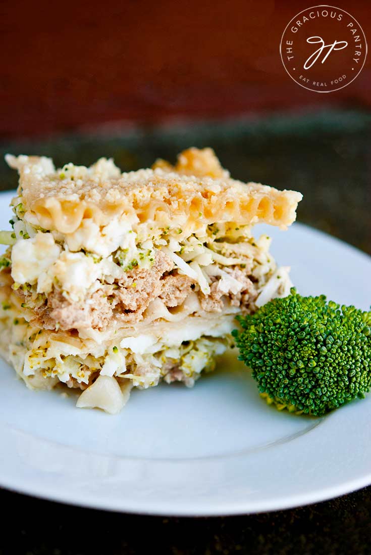 A slice of they Clean Eating White Lasagna Recipe sits on a white plate with a single broccoli floret beside it for garnish.