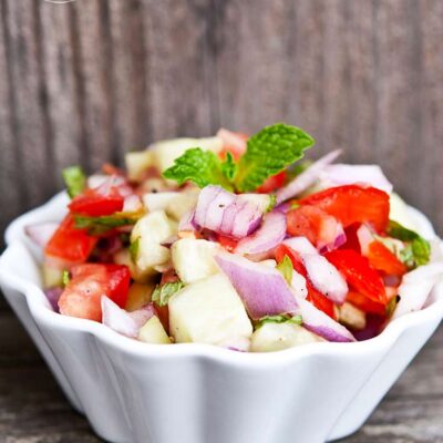 A white bowl sits on a wooden ledge, filled with this colorful Shirazi Salad.