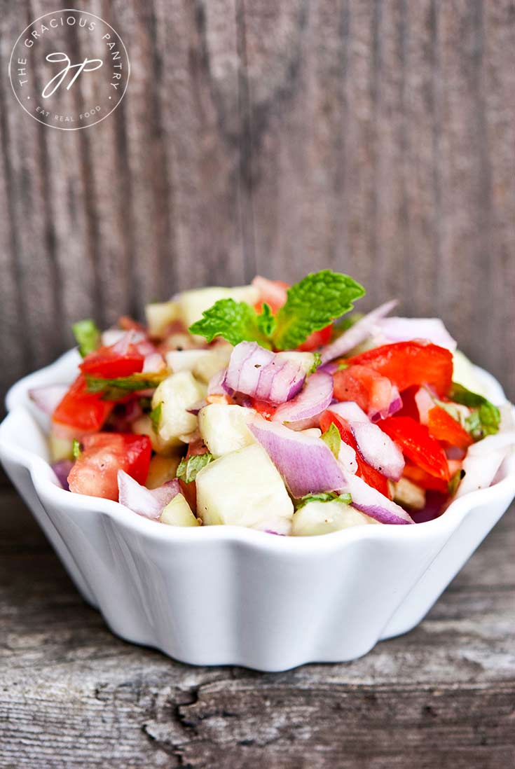 A white bowl sits on a wooden ledge, filled with this colorful Shirazi Salad.