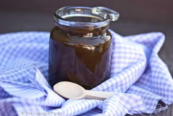 Showing the darker color of this sauce when made with a different sweetener. A dark caramel sauce fills a glass jar, a wooden spoon rests next to the jar.