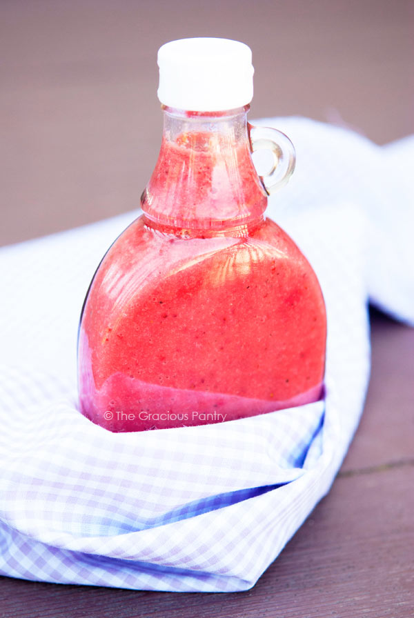 Clean Eating Strawberry Vinaigrette in a clear bottle showing it's bright, pink color. The bottle site nestled in a light purple and white checkered cloth.