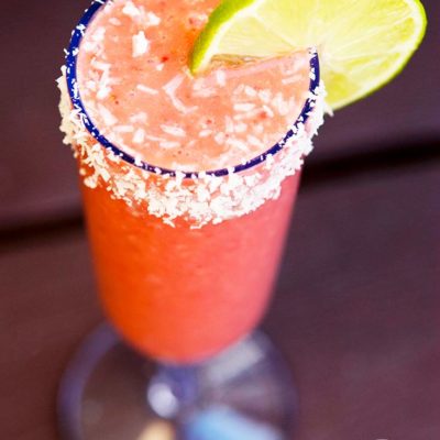 A delicious, strawberry daigquiri smoothie in a clear champagne glass, rimmed in coconut and garnished with a lime slice.