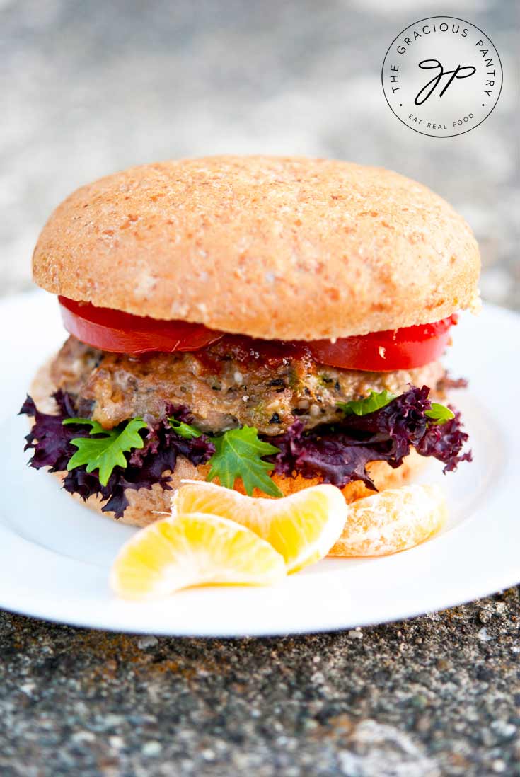 One of five Clean Eating Asian Style Portobello Mushroom Turkey Burgers sits on a white plate. You can see each layer from the bun to the meat patty, lettuce and tomato slices. In front of the burger on the rim of the plate sit a few mandarin orange slices as garnish.