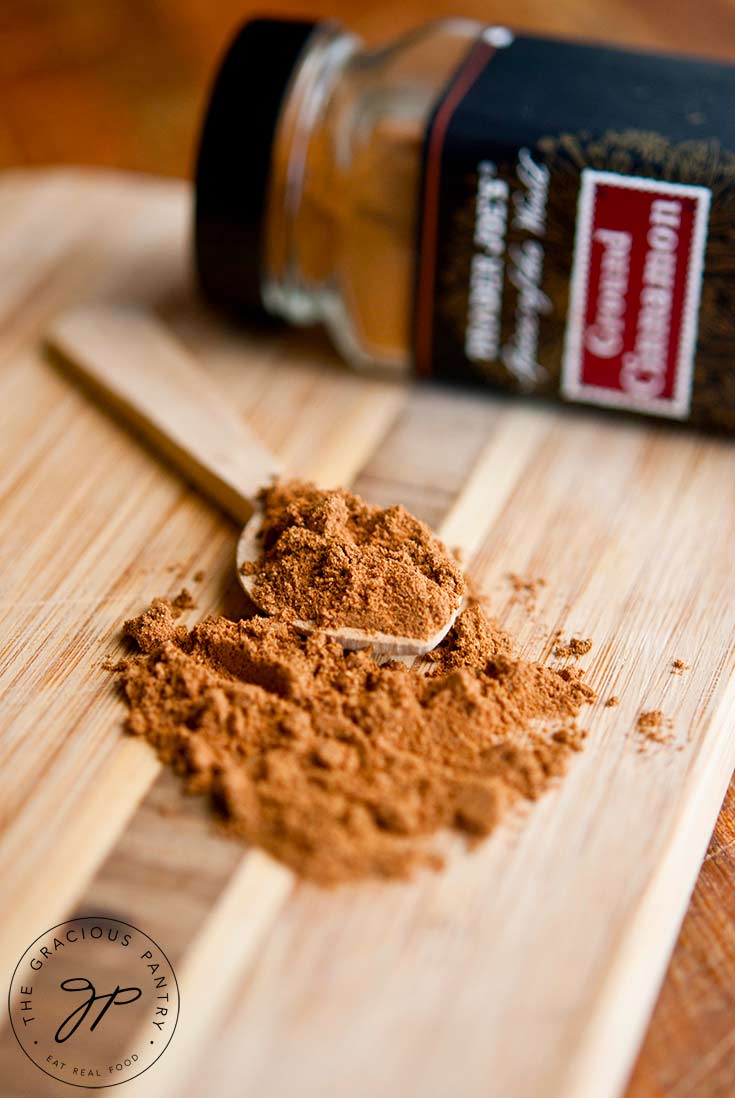 Some of this Pumpkin Pie Spice Recipe lays on a cutting board with a small wooden spoon ready to scoop some up. A bottle of cinnamon lays on it's side in the background.