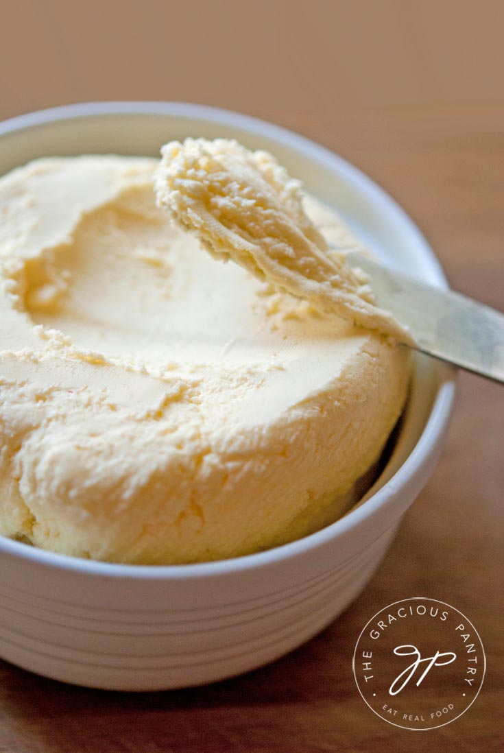 A close up shot of a white bowl holding freshly made, clean eating butter. A knife rests on the side of the bowl and has some butter scooped onto it.