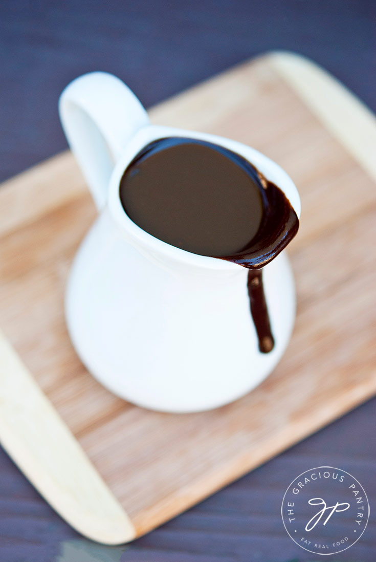 A white pitcher filled with this Clean Eating Coconut Fudge Sauce sits on a small cutting board as a bit of the fudge sauce runs down the front of the pitcher.