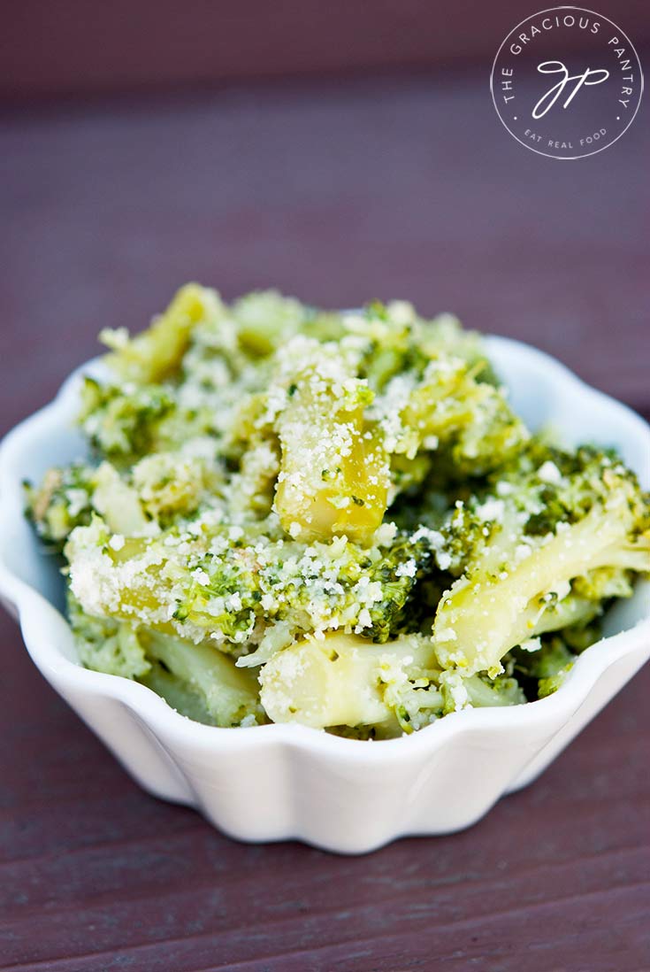 A white bowl sits filled with this Garlic Broccoli Recipe. You can see the specs of white parmesan coating the bright green broccoli florets. 
