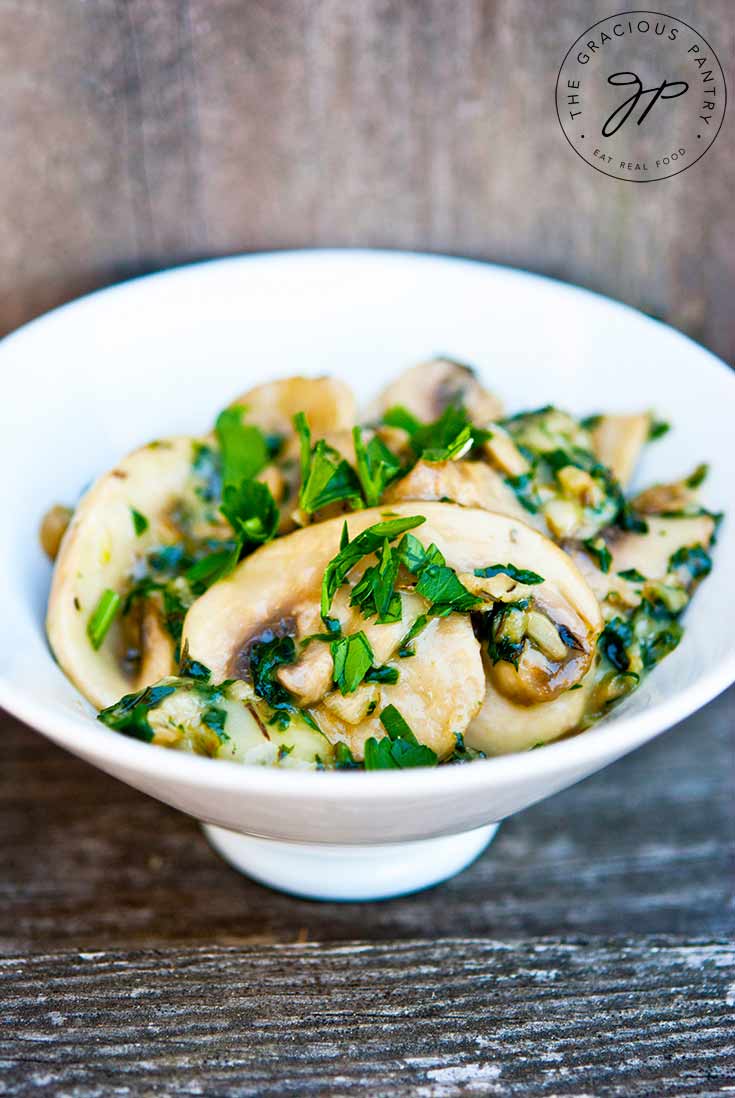 A single white bowl filled with this Clean Eating Garlic Mushrooms With Double Cream Brie Cheese sits on a wooden plank, ready to eat. You can see sprinkled of fresh parsley over the cheesy mushrooms.