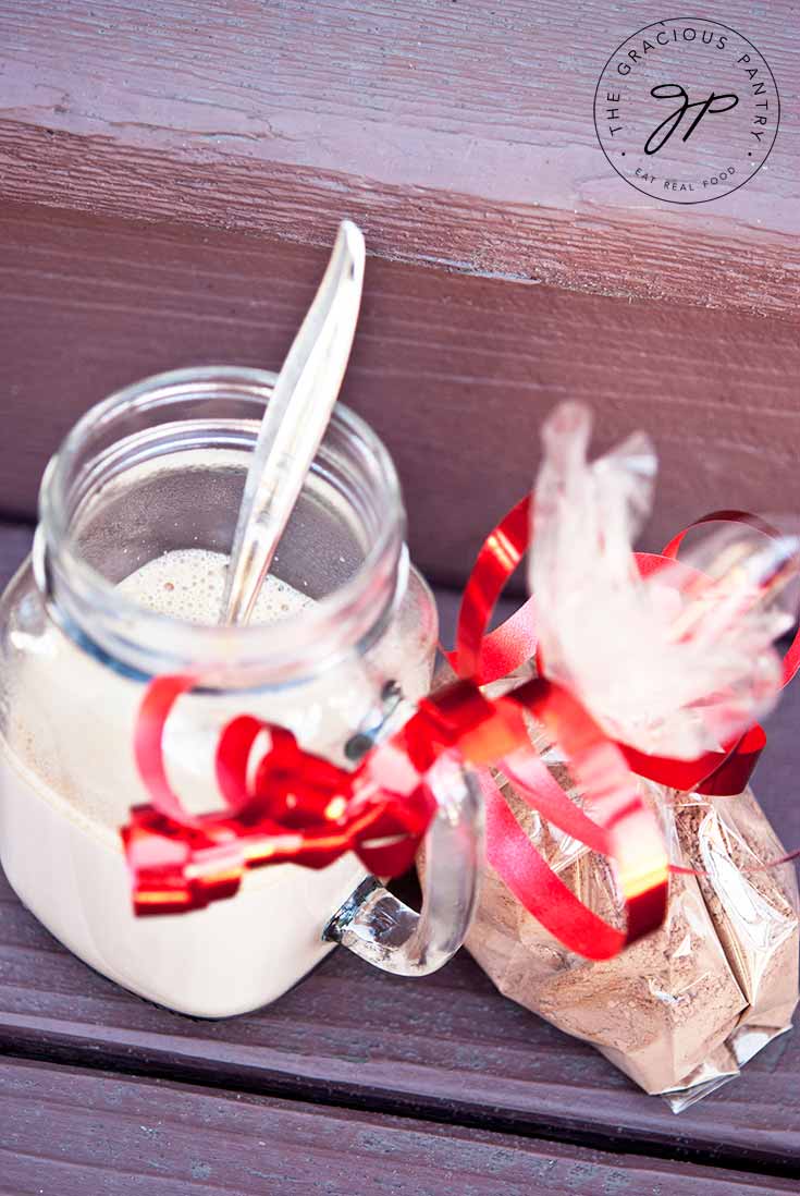 A mason jar mug sits on a wooden background filled with hot chocolate made from this Clean Eating Hot Chocolate Mix. A clear bag sits next to it filled with the dry mix and tied with a red ribbon.