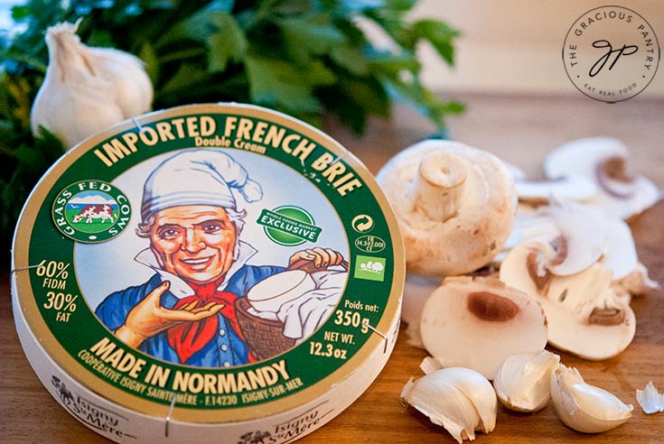 This shows the ingredients in this Clean Eating Garlic Mushrooms With Double Cream Brie Cheese. A wheel of brie in it's packaging sits next to some fresh, chopped parsley and raw mushrooms.