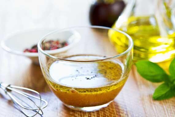 Image shows a clear glass cup with a small amount of oil-based salad dressing in it representing this information on Clean Eating Oils. Surrounding the glass are a small whisk, a larger bottle of oil and a fresh bunch of basil.
