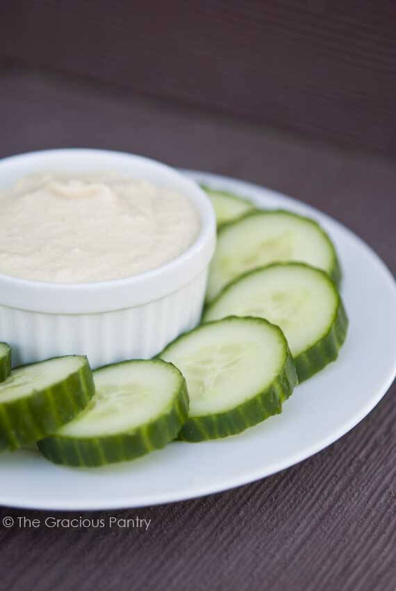 Clean Eating Tahini Sauce in a white bowl sitting on a plate, surrounded by sliced cucumbers for dipping.