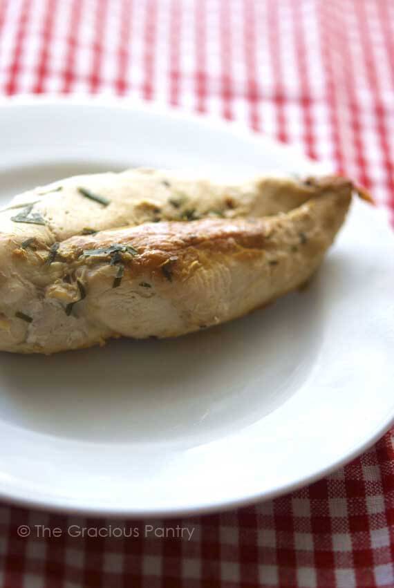 One of 8 Clean Eating Baked Chicken Recipes is sitting on a white plate. The chicken breast is baked and covered in herbs. The plate sits on a red and white checkered tablecloth.