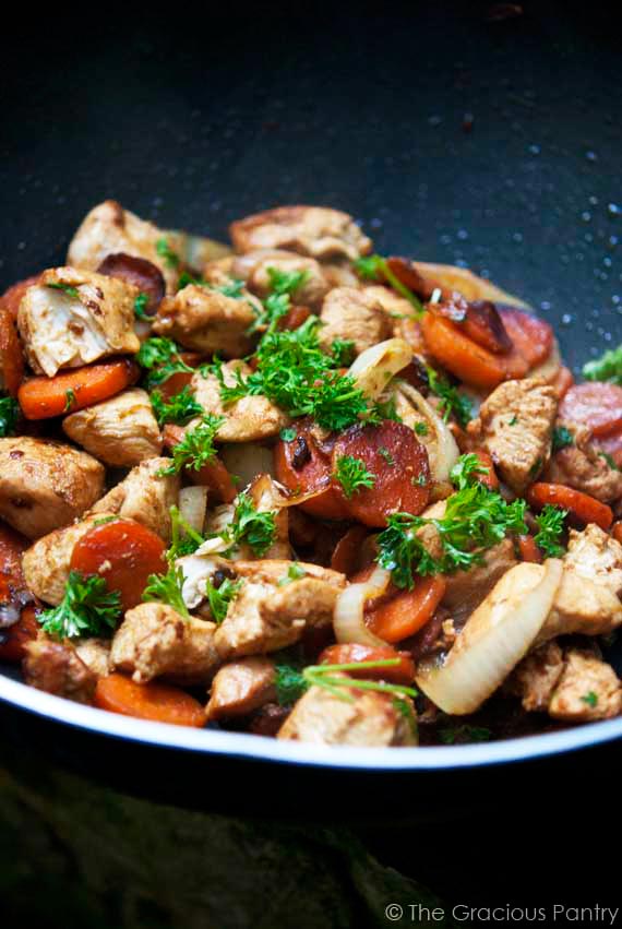 Clean Eating Wok-Style Garlic Chicken & Carrots recipe sitting in a black skillet with vibrant orange carrots and delicious looking chicken, sprinkled with fresh parsley for garnish.