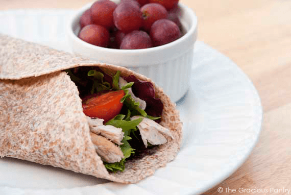 A Turkey Wrap on a white plate with a small bowl of red grapes.