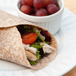 A single Turkey Wrap on a white plate next to a small white bowl of red grapes.