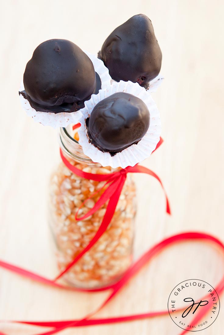 A jar filled with dry popcorn kernels holds three Clean Eating Chocolate Covered Strawberries on sticks. A red ribbon is tied around the jar and the ribbon falls nicely around the jar on the table.