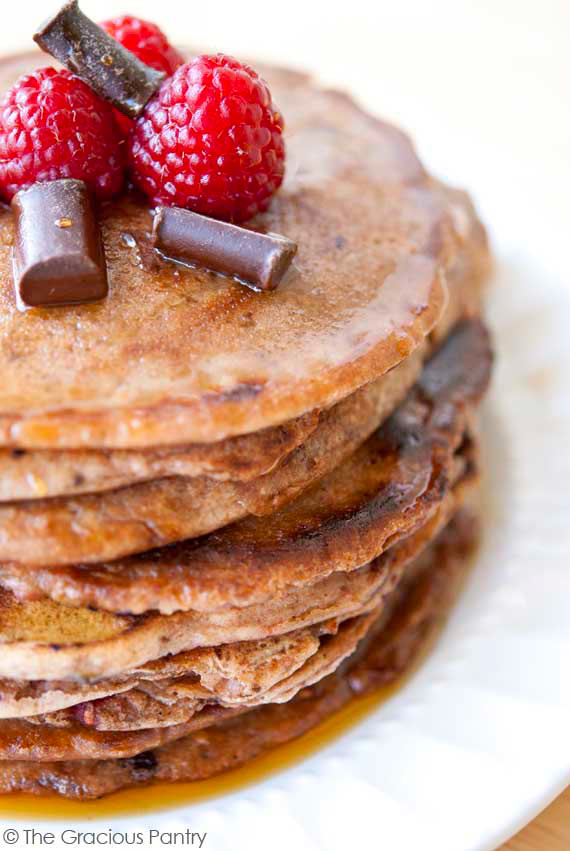 A stack of Clean Eating Raspberry Dark Chocolate Chip Pancakes sit stacked high on a white plate. You see a closeup of the pancakes, with the raspberries and the chocolate chips sitting on top of them.