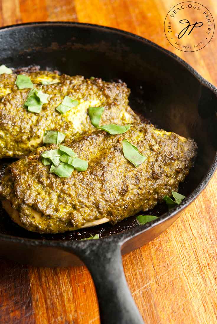 A black, cast iron skillet sits with two chicken breasts covered in pesto sauce and fresh basil sprinkled over the top in this Baked Pesto Chicken Recipe.