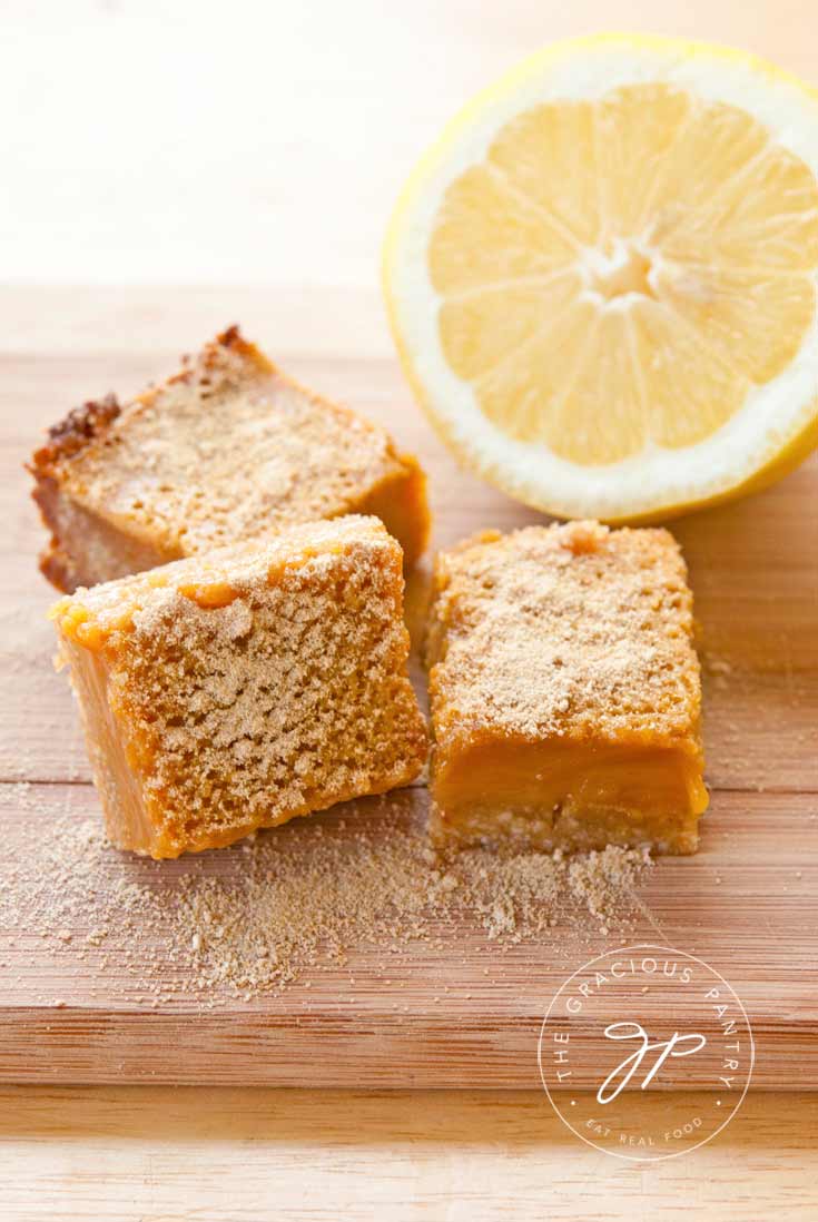 Three of these Clean Eating Lemon Bars sit on a wooden cutting board with half a lemon sitting next to them.