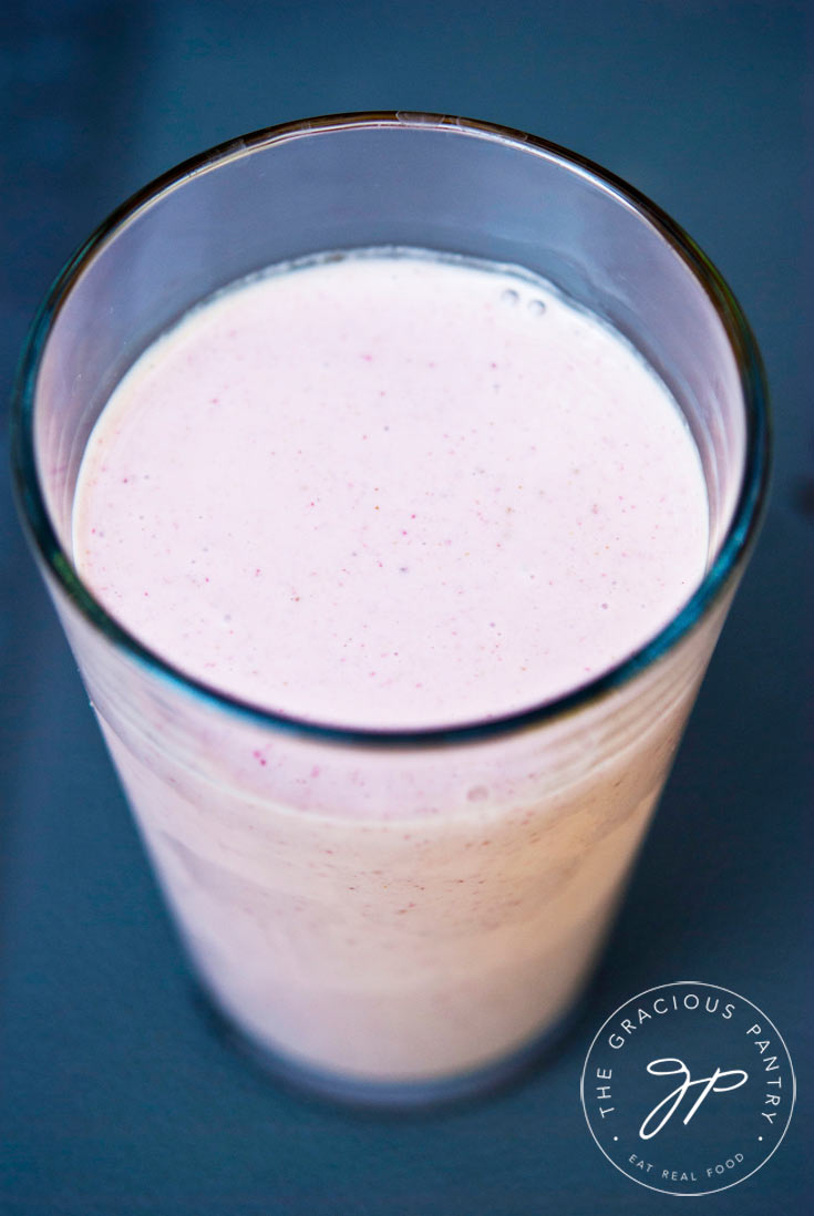 A tall, clear glass sits filled with Clean Eating Peanut Butter Strawberry Smoothie against a blue-gray background.